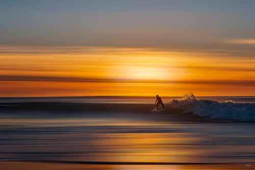 Photographie prise à la pointe du Cap-Ferret côté océan atlantique représentant la silhouette d'un homme surfant une vague au soleil couchant. La photo est très minimaliste et dynamique
