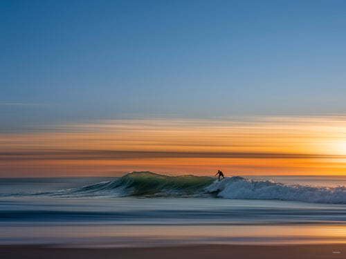 Photographie prise à la pointe du Cap-Ferret côté océan atlantique représentant la silhouette d'un homme surfant une vague au soleil couchant. La photo est très minimaliste et dynamique